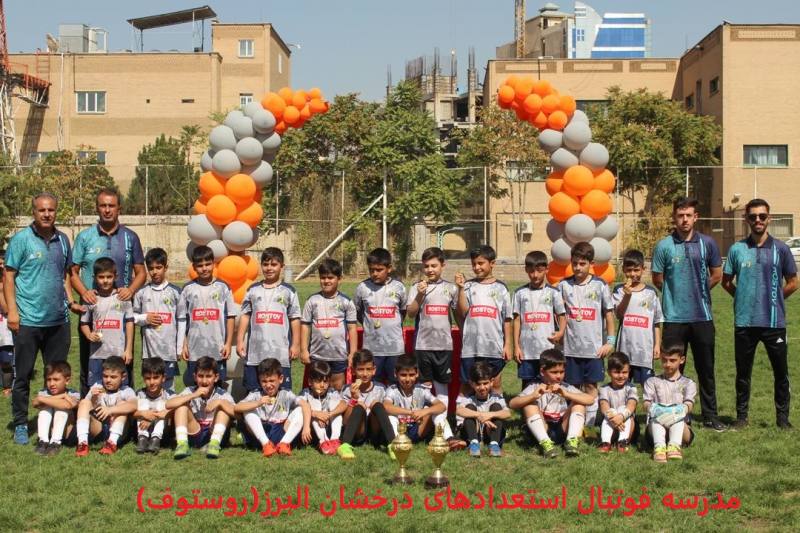 عکس تیمی فوتبالیستهای نونهال مدرسه روستوف کرج در مقابل تاج بادکنک با لباسهای طوسی و مدال در گردن در زمین چمن به همراه مربیان با لباس آبی