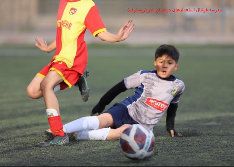 تصویر هیجان انگیز از بازی فوتبال دو نوجوان یکی با لباس طوسی و آرم روستوف و دیگری با لباس قرمز در زمین چمن